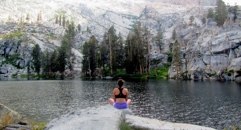 A person sits on a rock facing an alpine lake that appears to be surrounded by mountainous terrain and evergreen trees